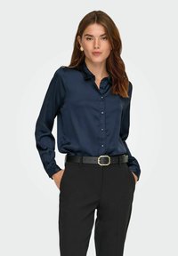 Marineblauwe satijnen blouse met een knoopsluiting, lange mouwen en een puntkraag, gecombineerd met zwarte broek en een zwarte riem met een gouden gesp.