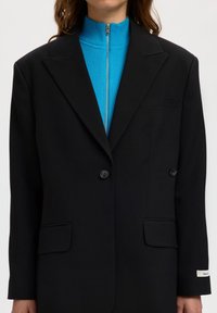 Γυναίκα με μαύρο σακάκι διπλού κουμπώματος πάνω από μπλε μπλούζα με φερμουάρ και ψηλό γιακά, τα χέρια χαλαρά δίπλα στο σώμα.
