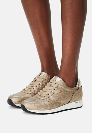 Goldfarbene Sneakers mit Struktur, Schnürdesign, metallischen Akzenten und kontrastierender schwarzer Sohle. Auffälliges Logo-Detail an der Seite.