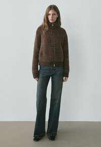 Brauner, strukturierter Reißverschluss-Cardigan mit hohem Kragen und großen Fronttaschen, kombiniert mit ausgestellten, dunkelblauen Jeans.