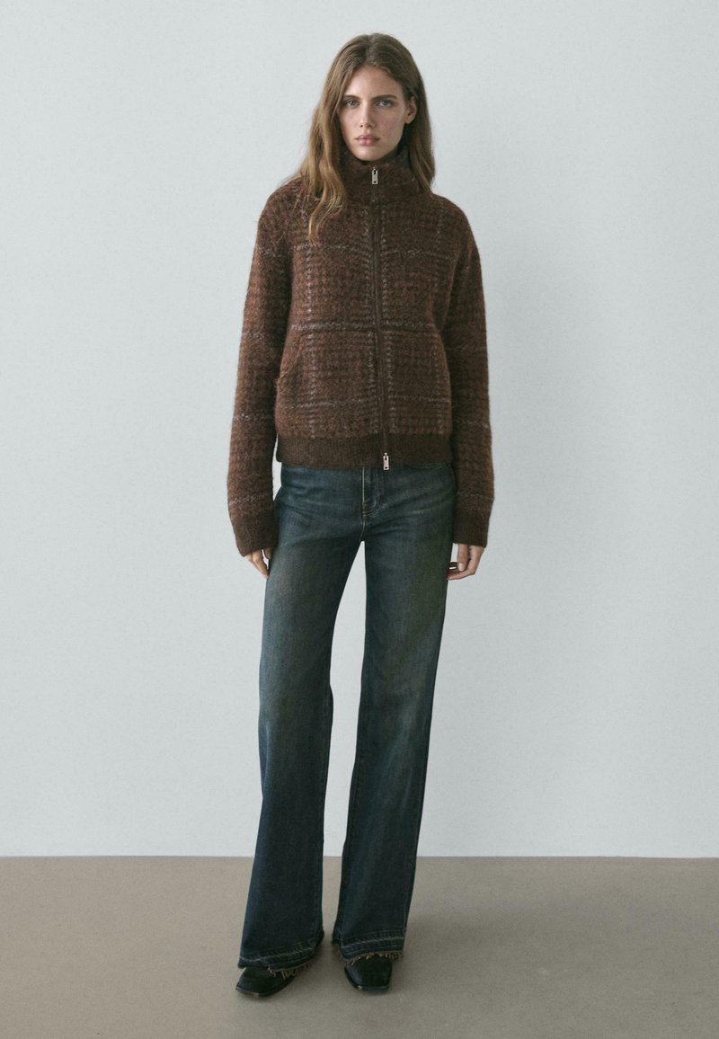 Brązowy sweter na zamek z wysokim kołnierzem i dużymi przednimi kieszeniami, w połączeniu z rozszerzanymi ciemnoniebieskimi dżinsami.