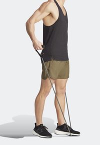 Débardeur de sport noir, short vert olive et baskets noires. Personne tenant des bandes de résistance pour l'exercice, mettant en valeur une silhouette musclée.