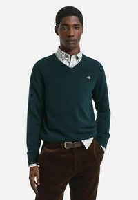Dunkelgrüner V-Ausschnitt-Pullover aus weichem Material, über ein leicht kariertes Hemd getragen. Braune Cordhosen mit einem braunen Gürtel.