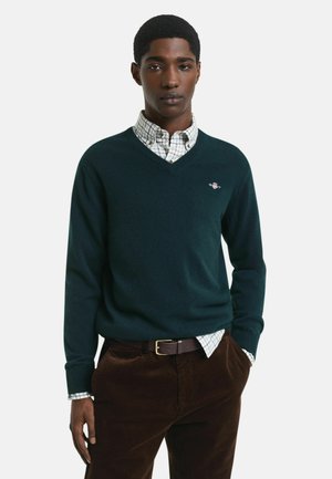 Dunkelgrüner V-Ausschnitt-Pullover aus weichem Material, über ein leicht kariertes Hemd getragen. Braune Cordhosen mit einem braunen Gürtel.