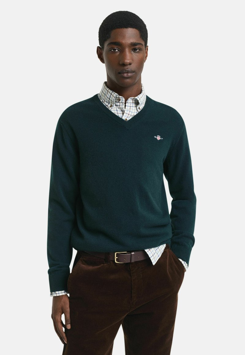 Dunkelgrüner V-Ausschnitt-Pullover aus weichem Material, über ein leicht kariertes Hemd getragen. Braune Cordhosen mit einem braunen Gürtel.