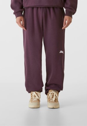 Lila Jogginghose mit einem elastischen Bund und Bündchen, ausgestattet mit einem kleinen weißen Logo am linken Hüftbereich. Kombiniert mit hellbeigen Sneakern.