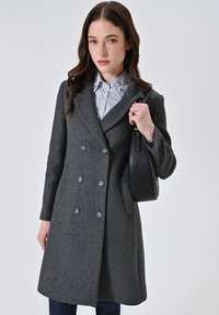 Grau melierter Wollmantel mit doppelter Knopfleiste, tailliertem Schnitt und Seitentaschen. Kombiniert mit einem gestreiften Hemd und einer schwarzen Handtasche.