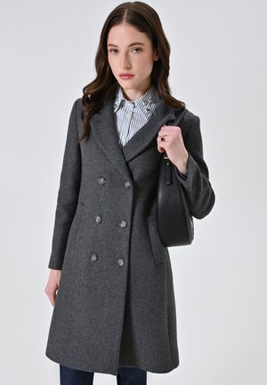 Cappotto grigio in misto lana con chiusura a doppio petto, vestibilità sartoriale e tasche laterali. Abbinato a una camicia a righe e una borsa nera.