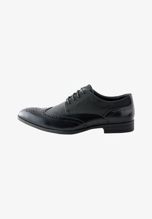 Black men's leather oxford shoe with wingtip brogue detailing and fabric panels, laced, with a low heel and pointed toe.