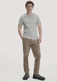 Jonge man in een grijze T-shirt met korte mouwen, lichtbruine corduroy broek en twee-tint veterschoenen, staande met één hand in zijn zak.