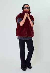 Burgundinė dirbtinė kailio liemenė su aukštu apykakle, dėvima virš pilkos palaidinės. Modelis vilki tamsiai mėlynas vertikalių dryžių kelnes ir tamsius batus, papildytas mėlynomis saulės akimis.