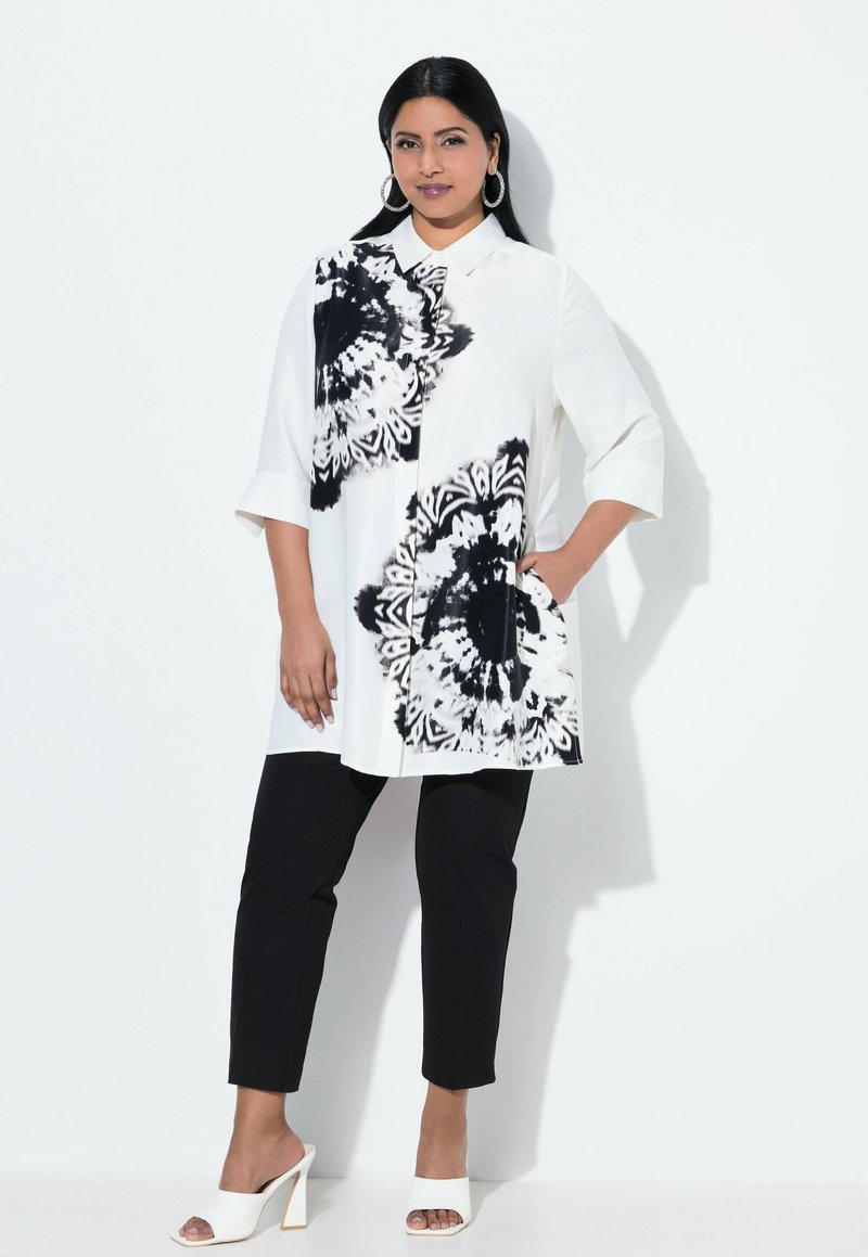 White tunic with black floral design, short sleeves, collar, and side pocket, paired with black slim-fit pants and white open-toe heels.