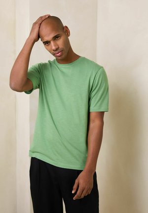 Mann in grünem T-Shirt und schwarzer Hose berührt seinen kahlen Kopf, steht vor einem schlichten beigen Hintergrund mit nachdenklichem Gesichtsausdruck.