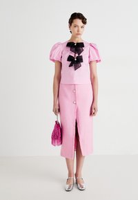 Kvinne som har på seg en rosa topp med puffermer og svarte sløyfer, et rosa midi-skjørt med knapper, sølvfargede flate sko, og bærer en rosa veske med frynser.