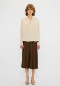Beige Bluse mit langen Ärmeln und V-Ausschnitt, kombiniert mit einem braunen, plissierten Midirock und beige spitz zulaufenden Ballerinas mit dekorativen Akzenten.