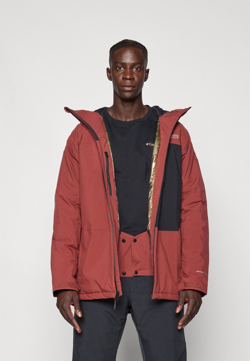 Giacca impermeabile rossa con cappuccio, caratterizzata da accenti neri, una cerniera dorata e un design imbottito. Indossata sopra una maglietta nera.