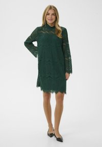 Zelené krajkové šaty s dlouhými průhlednými rukávy, vysokým výstřihem, vlnitým lemem a složitými geometrickými vzory. Nosí se s černými botami s špičatou špičkou.