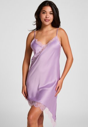 Jeune femme portant une nuisette en satin lavande sans manches avec bordure en dentelle, debout devant un fond uni gris clair.