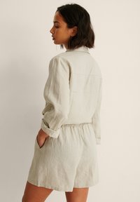 Light gray linen romper with a collared neckline, long sleeves rolled up, elastic waist, and side pockets. Smooth, slightly textured fabric.