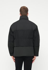 Juodas pūkinis švarkas su aukštu apykakle, turintis segmentuotą dizainą su horizontaliomis juostelėmis, pagamintas iš lygaus, matinio audinio.