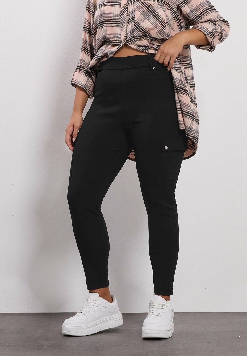 Leggings cargo neri elasticizzati con tasche laterali e una texture liscia, abbinati a una camicia a scacchi rosa e nera e sneakers bianche.