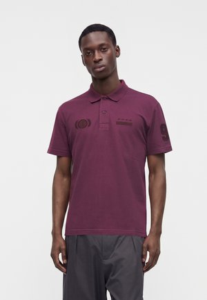Jeune homme portant un polo bordeaux à manches courtes avec des détails brodés et un pantalon gris foncé, debout devant un fond uni.