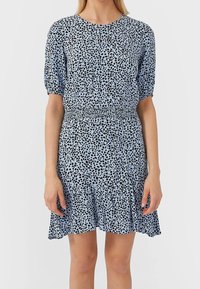 Blå klänning med svarta prickar, korta pufsig ärmar, smockad midja och volangkant. Lättviktig tyg med en jämn textur.