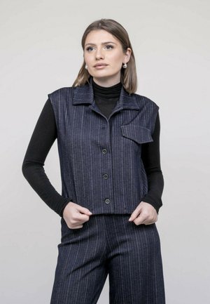 Femme portant un gilet sans manches à fines rayures sombres sur un haut noir à manches longues et un pantalon assorti à fines rayures, devant un fond uni.