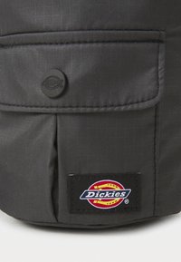Zwarte stoffen tas met een gestructureerd oppervlak, een voorzak, een ronde zwarte knop en een kleurrijke Dickies logo patch.