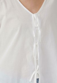 Top in lino bianco con scollo a V, dotato di lacci sul davanti per la chiusura e una leggera texture. Design semplice e leggero.
