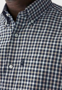 Camisa de cuadros azules y blancos con detalles en marrón, cuello abotonado y botones metálicos; presenta un logo bordado en el bolsillo del pecho.