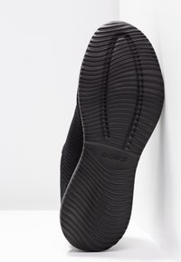 Semelle d'une chaussure noire BOBS montrant des rainures texturées et incurvées pour l'adhérence avec le nom de la marque embossé près du centre de la semelle.