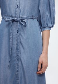Gros plan sur une robe en denim avec des manches bouffantes et une ceinture en tissu assorti nouée à la taille, portée par une personne dont la main droite est visible.