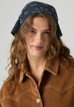 Jonge vrouw met lang golvend haar die een donkerblauw patroon hoofddoek en een bruine corduroy jas draagt, kijkt naar de camera.
