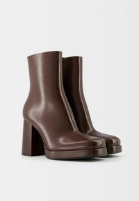 Hnědé kožené kotníkové boty s kulatou špičkou, robustním vysokým podpatkem a elegantním designem. Hladký povrch s minimálním zdobením.