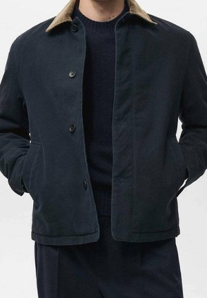 Homme portant une veste bleu marine foncé avec un col beige, partiellement boutonnée, mains dans les poches, par-dessus un pull sombre et un pantalon assorti.