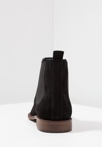 Bottine en daim noir avec panneaux latéraux élastiques, bout arrondi et talon en bois marron. Texture lisse avec un minimum de détails.