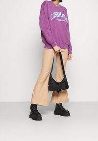 Fialová mikina s grafikou "COPENHAGEN", béžové rozšířené kalhoty a černé robustní šněrovací boty. Černá taška držena jedním popruhem.