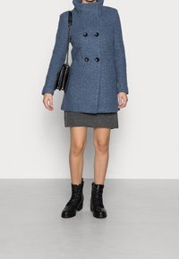 Modrý kabát z vlněné směsi s dvouřadým zapínáním, černými knoflíky a vysokým límcem, v kombinaci se šedou sukní a černými kotníkovými botami na šněrování.