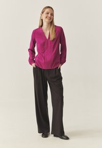 Donna sorridente con lunghi capelli biondi, indossa orecchini d'oro, una blusa magenta, pantaloni neri larghi e scarpe nere, mentre sta in piedi con le mani in tasca.