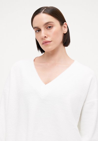 White sweater with a V-neck, made of textured fabric, featuring wide sleeves and a relaxed fit, set against a light background.