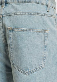 Lichtblauwe denim jeans hebben een achterzak met een piekvormig ontwerp. De stof heeft een subtiele textuur en zichtbare stiksels.