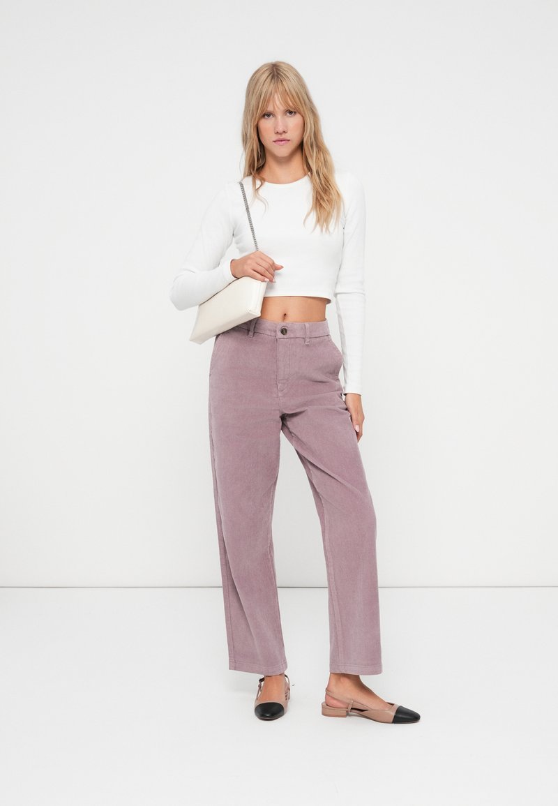 Pantalones de pana anchos en color lila claro con bolsillos frontales y cierre de botón único, combinados con un top corto de manga larga blanco y zapatos planos bicolores.