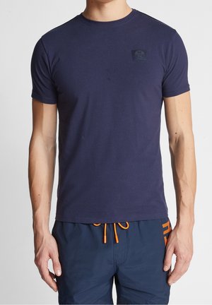 Mężczyzna w dopasowanym granatowym t-shircie z krótkim rękawem i granatowych szortach z pomarańczowym sznurkiem i napisem na boku.