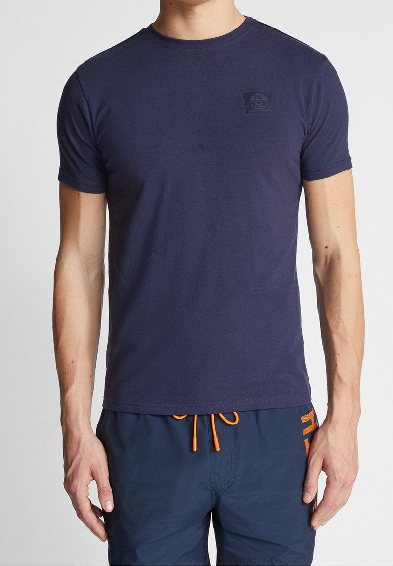 Homme portant un t-shirt bleu marine ajusté à manches courtes et un short bleu marine avec un cordon orange et un texte sur le côté.