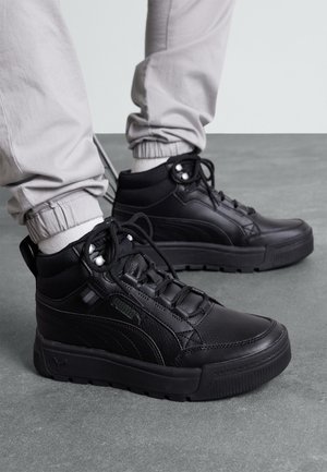 Czarne sneakersy za kostkę z grubymi podeszwami noszone z jasnoszarymi spodniami dresowymi na szarej podłodze.