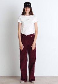 Bílá bavlněná tričko s potiskem, doplněné širokými burgundy sametovými kalhotami. Model nosí černou kšiltovku. Hladká textura, uvolněný střih.