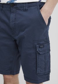 Pantaloni scurți cargo de culoare albastru marin, fabricați dintr-un material ușor, având buzunare laterale, detalii de cusătură și un patch cu text pe piciorul drept.
