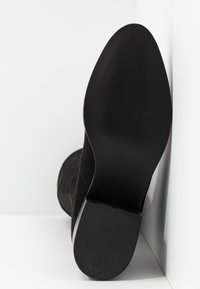 Botín de ante negro con suela de goma texturizada, que presenta un diseño elegante y una punta redondeada, mostrado desde la perspectiva del talón.