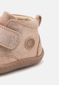 Botines de ante rosa con una correa de velcro. Forma redondeada, acabado texturizado y suela de goma prominente. Presenta un detalle de logo circular.